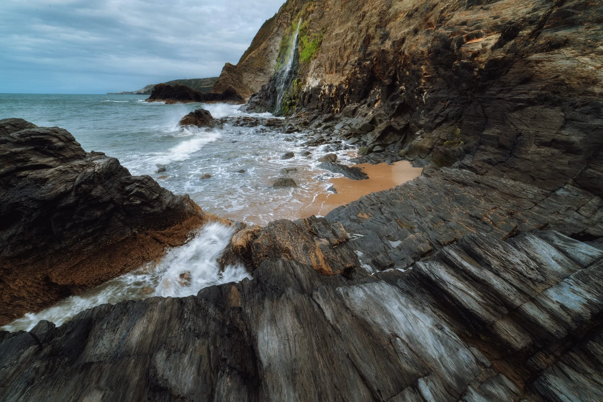  The northern cliffs of Tresaith are more popular, and this waterfall plunging into the sea is the main reason why. After some clambering over and around the slippery rocks, I lined up several compositions involving the incredible crag geology, the cliffs, and the waterfall. This one ended up being my favourite because of the extra energy lent by the rushing waves. 