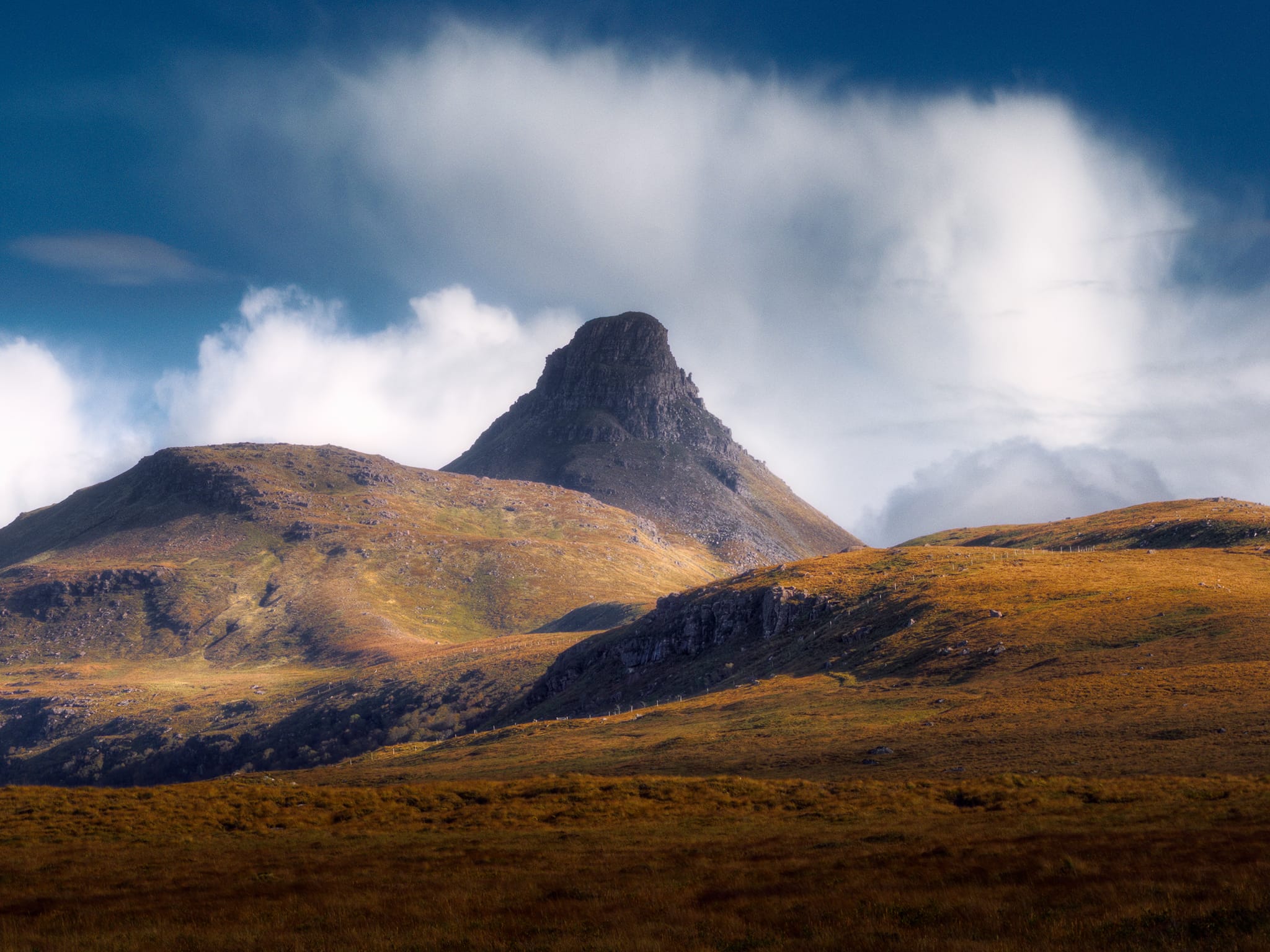 Further along the road the clouds were finally starting to move on. We drove past Loch Lurgainn then Loch Bad a’ Ghaill, taking the road at Badnagyle up and onto the moorland between Stac Pollaidh and Loch Garvie. It was here that I got my first side-profile view of Stac Pollaidh, looking like a slender pyramid topped with crags and pinnacles. The clouds broke apart and raced away, allowing light to pick out the beautiful contours of the landscape as I lined up this zoomed in composition of what was quickly becoming one of my favourite mountains.