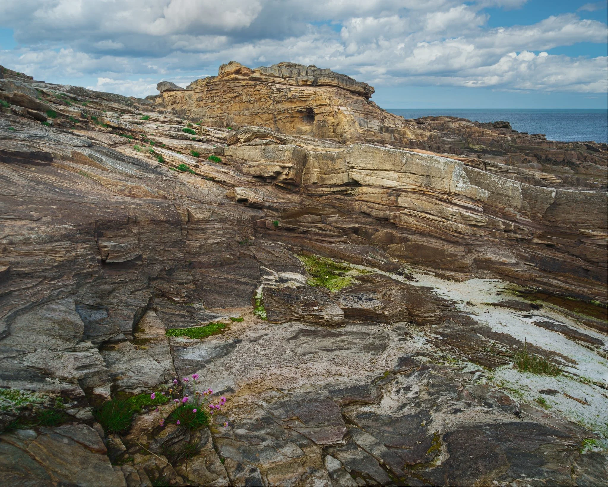  A fantastical mish-mash of geology here. In the foreground, smooth, low-angle siltstones/mudstones formed from old overbank (floodplain) mud settling out when the river crept over its banks. Modern tides, salt spray and lichens have bleached and smoothed those laminations, making them look chalky. The subtle, sharp break (you can just make it out sloping away) marks where a later channel scoured into those muds. The big yellow-buff blocks are the  Rumbling Kern  channel sandstones: coarse, cross-bedded braid-plain sand dumped by fast currents. 