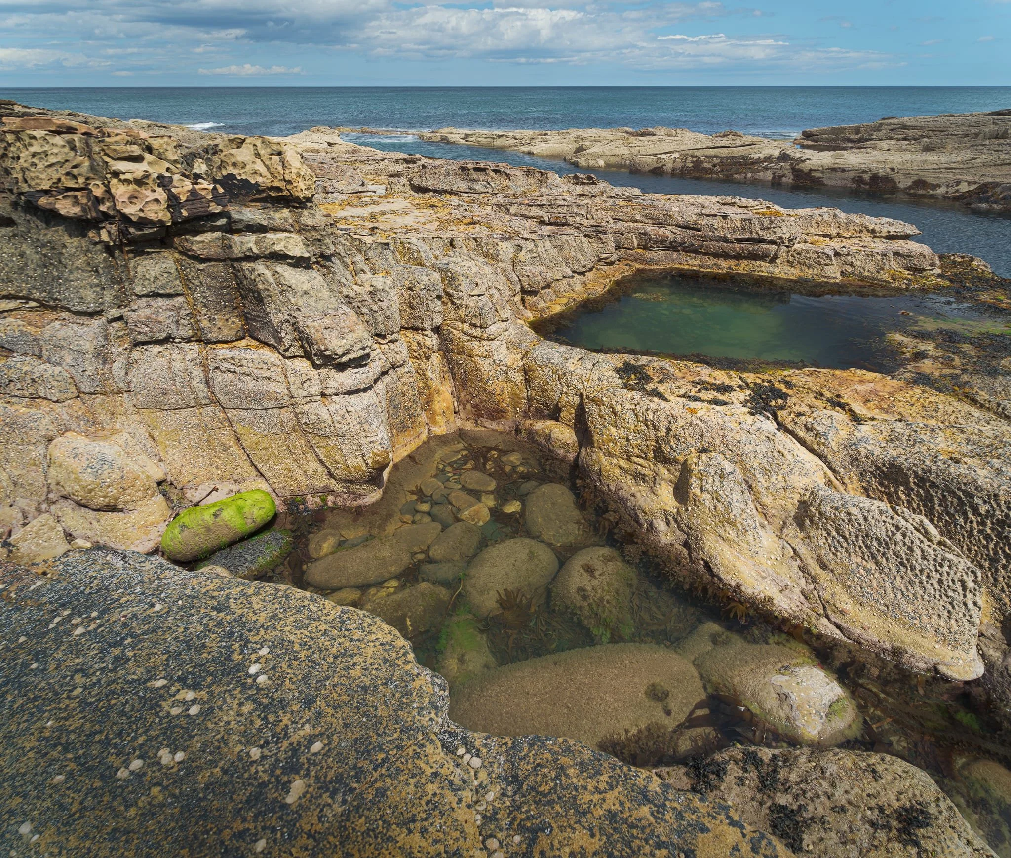  This shot was made from three landscape images at 24mm, stacked top to bottom. Small, circular depressions have been drilled into the sandstone by countless swirls of sand-and-pebble slurry at low tide. The lacy, pitted &ldquo;bee-nest&rdquo; pattern on the block tops comes from salt-crystal growth and repeated wet-dry cycles; tiny pockets where salt expands and flakes the rock. Vertical fractures and bedding planes guided where water could exploit weaknesses, prising blocks apart and focusing erosion at those joints and corners. 