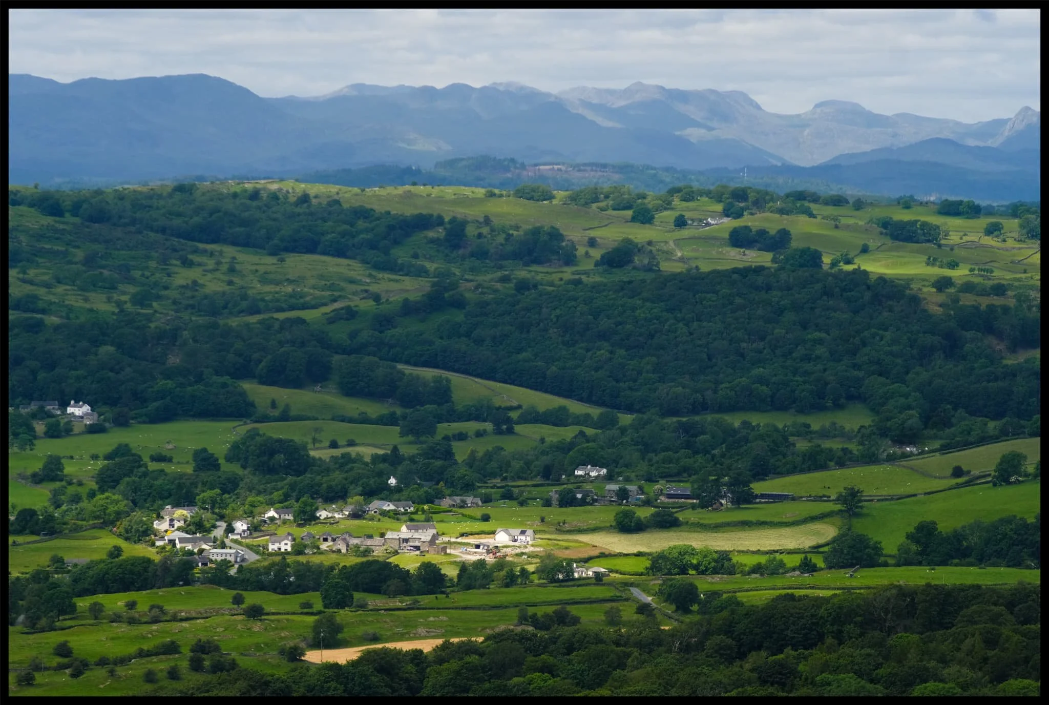  One of the Lyth Valley&rsquo;s little villages gets illuminated by the sun as it breaks through the clouds. The Lake District fells rise above. 