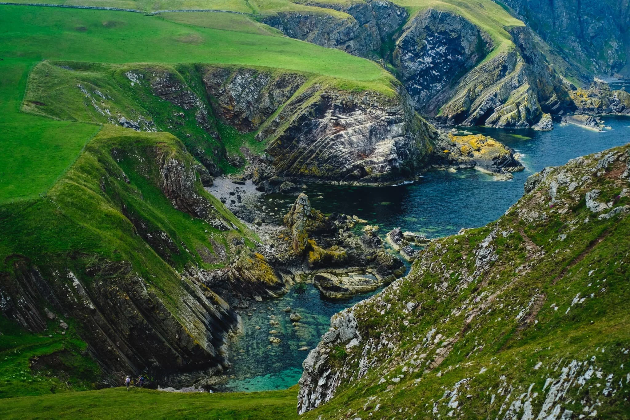  To get a better view of the Uilystrand Brae cliffs, we elect to follow the steep grassy path down to Pettico Wick Bay, with the views getting more epic along the way. Check out the wee little hikers in the bottom-left of the frame! 