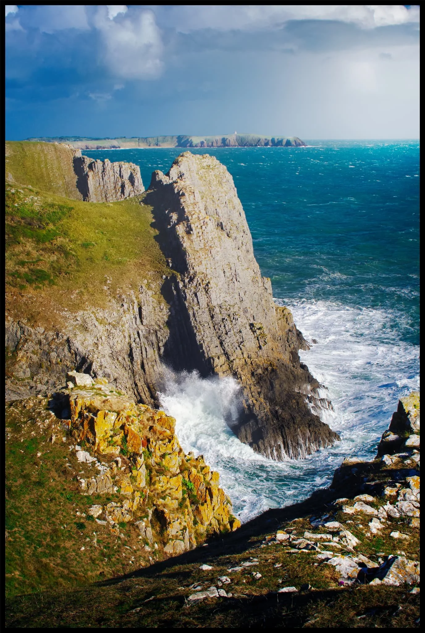  A slow-moving squall sits above Caldey Island in the distance whilst the sun drench the Lydstep cliffs with warm late-afternoon light as powerful winds blast the sea at the seemingly impossible cliffs. 